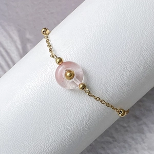 Macro view of gold chain bracelet with pink gemstone and beads on a white pedestal