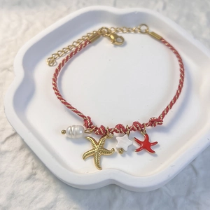Detail of red braided bracelet with golden starfish, pearl and coral charms on white tray