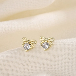 Elegant gold bow earrings with a central clear zircon stone on a beige textured fabric
