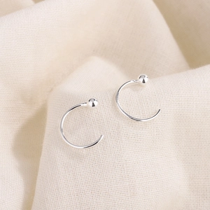 Minimalist C-shaped silver hoop earrings with smooth bead accents on beige fabric background