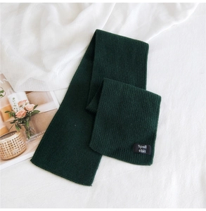 Detail of dark green ribbed knit scarf with woven brand label on soft white linen background