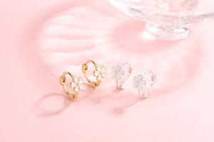 Close-up of gold and silver S925 floral earrings with white enamel petals on pink surface