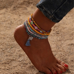 Colorful beaded bohemian anklets with a heart charm and tassel detail on beach sand