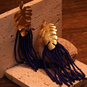 Detail of gold monstera leaf earrings with deep blue beaded tassels on stone background