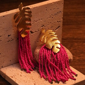 Detailed view of tropical leaf earrings with rose red beaded tassels on a stone stand