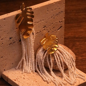 Detailed close-up of golden monstera leaf earrings with long white beaded tassels