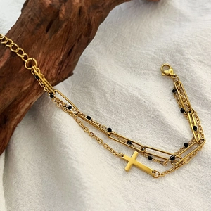 Double-layer gold cross bracelet with black beads and paperclip chain on wooden stand