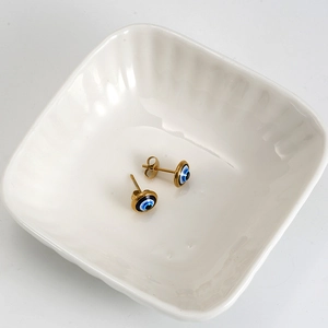Close-up of golden Evil Eye stud earrings with blue resin in a white ceramic dish