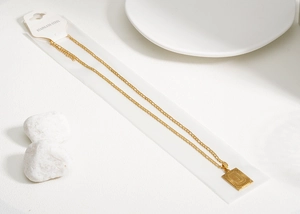 Détail macro du pendentif carré doré avec la lettre J sur une chaîne Figaro en acier