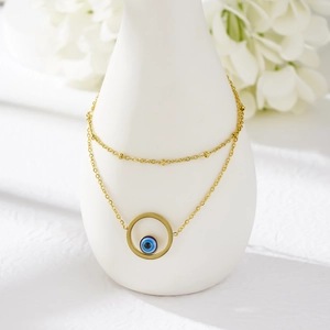 Gold double-layered stainless steel anklet with blue evil eye pendant on white ceramic