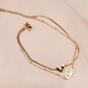Detail of gold double-layer anklet with heart charm and love pendant on soft fabric