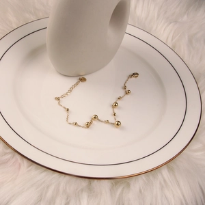 Close-up of golden bead anklet with lobster clasp on white porcelain tray in soft light