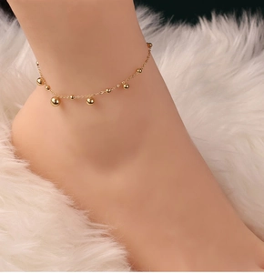 Macro shot of minimalist gold beaded anklet worn on ankle over soft white plush background