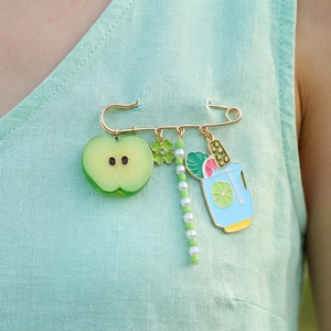Golden safety pin brooch featuring green apple, clover, and blue cocktail charms on linen