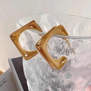 Modern geometric square gold hoop earrings displayed on an artistic glass cup rim