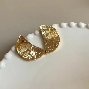 Macro shot of gold fan-shaped stud earrings with sunburst texture on white ceramic tray