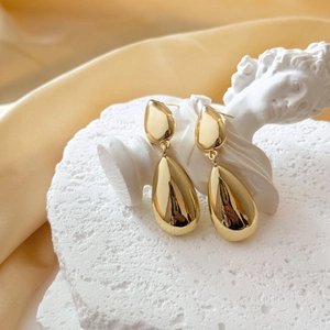 Polished gold double teardrop earrings displayed on a white stone pedestal with silk