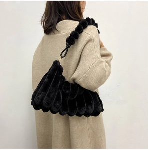 Fashionable black plush shoulder bag featuring a unique pom-pom strap worn on a beige knit sweater
