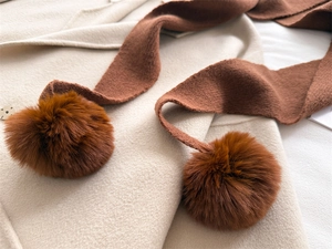 Détail d'une écharpe en maille caramel avec pompons en fourrure sur manteau blanc