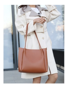 Caramel leather tote bag with subtle side pleats shown on a model in a trench coat
