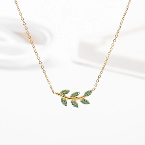 Détail d'un collier feuille dorée avec zircons verts sur fond blanc minimaliste