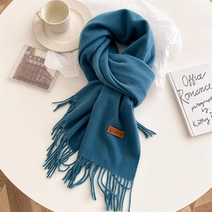 Close-up of deep blue knit scarf with fringe trim and leather brand label on white table