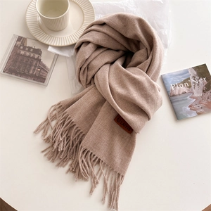 Detail of camel wool knit scarf showing fringe edge and leather logo on white desk