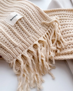 Macro shot of off-white knitted wool scarf with wavy ribs, tassels, and a woven brand label