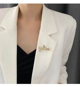 Detailed view of a gold mermaid tail brooch with crystals and a white pearl on a white lapel