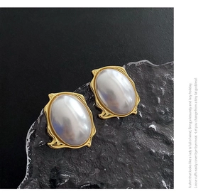 Detail of oval pearl earrings with gold wavy frames on a textured black backdrop