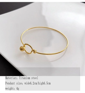 Close-up of golden titanium steel bangle with ball charm and hook clasp on white ceramic
