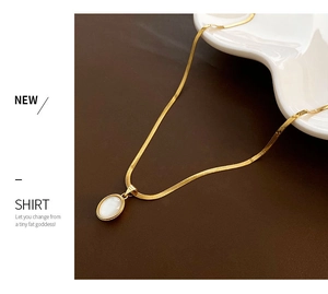 Macro detail of gold snake chain with white oval shell pendant on dark brown background
