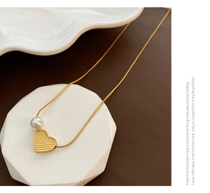 Detail of gold snake chain necklace with pearl and engraved heart charm on white stone