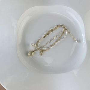 Detail of layered gold bracelet with white pearls and heart pendant on white ceramic plate