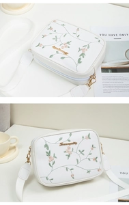 Detail of floral embroidery and gold logo on white crossbody camera bag with wide strap