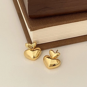 Detail showing stacked golden 3D heart stud earrings on a minimalist cream background