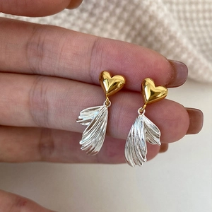 Asymmetrical gold heart and silver wing dangle earrings held for scale demonstration