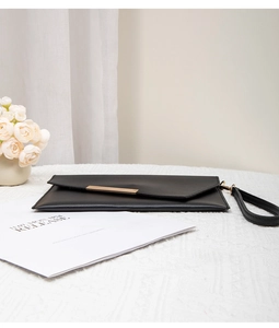 Detail of black envelope clutch bag with gold metal trim and wristlet on white table