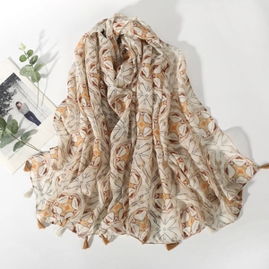 Écharpe beige à motifs géométriques et pompons sur fond blanc avec feuilles décoratives