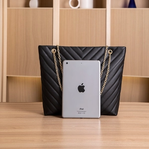 Macro view of black V-quilted tote bag with gold chain strap next to an iPad for size