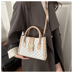 Beige tote bag with grey M-letter print and gold metal logo shown on model's outfit