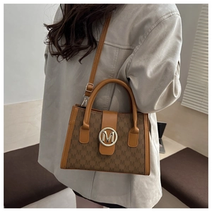 Close-up of tan monogram shoulder bag with gold 'M' buckle and structured trapezoid shape