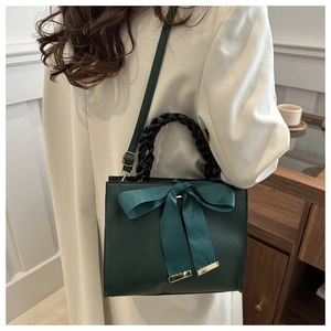 Sac à main carré vert foncé avec grand nœud et poignée tissée noire porté par un modèle