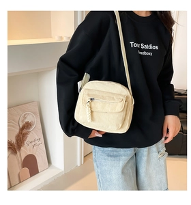 Detail of a cream-white corduroy crossbody bag with woven strap and decorative pendant