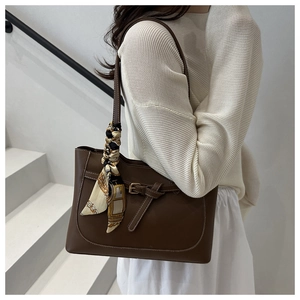 Brown leather tote bag with decorative silk scarf on handle held by a fashion model