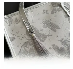 Macro detail of silver tote bag featuring butterfly embroidery and grey tassel charm