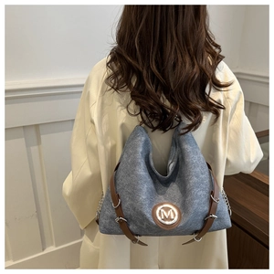 Détail du cabas gris-bleu montrant le logo M en métal et la sangle en cuir marron