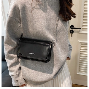 Detail of a black patent leather crossbody bag with silver chain strap worn by a model