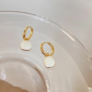 Detail of elegant gold square earrings hanging on a glass cup with cream enamel finish