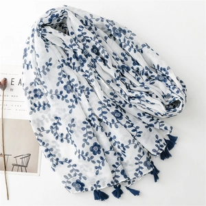 White linen scarf with blue floral embroidery and tassels shown in a natural setting